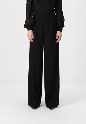 Zwarte wijde broek met een gladde textuur, hoge taille en aangepaste plooien. Heeft spitse zwarte hakken. Minimalistisch ontwerp.