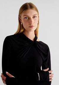 Top noir à manches longues avec un détail de nœud tordu au niveau du col et des accentuations sans manches aux poignets, fabriqué dans un tissu lisse et extensible.