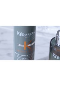 Kérastase Genesis Homme sprayflaska, cylindrisk form, matt mörkgrön med silverdetaljer och orange text, som innehåller produktinformation.