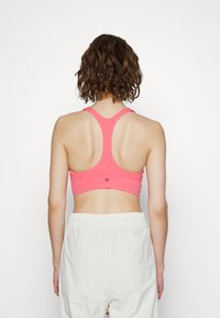 Sutien desportivo rosa com um design de costas nadador, textura suave, alças finas e um pequeno detalhe de logo nas costas. Combinado com calças de cor clara.