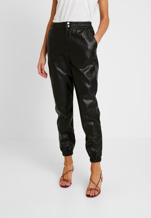 Femme portant un pantalon de jogging noir en similicuir avec des chevilles et une taille élastiques, associé à un haut sans manches blanc et des sandales à brides rouges.
