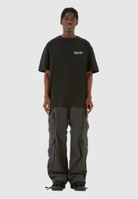 Tricou oversize negru cu logo albastru și pantaloni cargo gri, cu multiple buzunare și croială relaxată, asortați cu adidași negri.