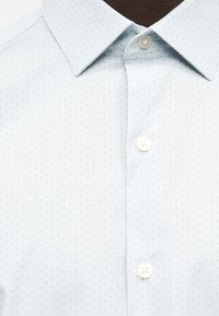 Λευκό πουκάμισο με μικρό σχέδιο από ανοιχτό μπλε τελείες, μυτερό γιακά και τρία ορατά λευκά κουμπιά στο μπροστινό μέρος.