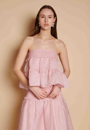 Jeune femme portant un haut rose texturé sans bretelles et une jupe assortie, debout devant un fond beige, mains jointes à la taille.