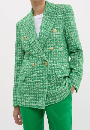 Frau, die einen grünen, strukturierten, doppelreihigen Blazer mit goldenen Knöpfen und passende grüne Hose trägt, die Hand in der Tasche.