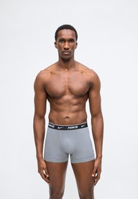 Μυώδης άνδρας στέκεται αντικρίζοντας μπροστά φορώντας γκρι εσώρουχα-μπόξερ Nike με μαύρη μέση, σε απλό λευκό φόντο.