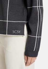 Ciemnoszary sweter z białym wzorem w kratę, z bocznymi rozcięciami i logo "SCSY" na dole. Dzianinowa tekstura i ściągacze przy mankietach.