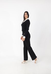 Suéter negro de canalé con cuello de volantes blanco y botones, combinado con pantalones de campana negros y sandalias de tacón crema.