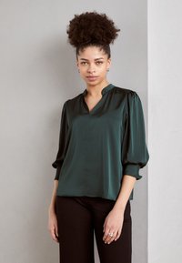 Blouse en satin vert émeraude avec un plastron ton sur ton, des manches bouffantes et une coupe décontractée, associée à un pantalon noir et des bagues en argent.