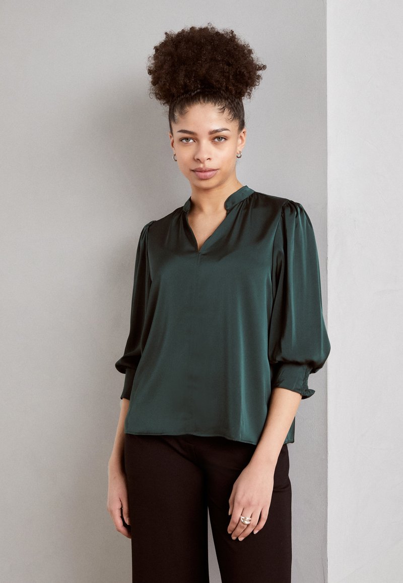 Blouse en satin vert émeraude avec un plastron ton sur ton, des manches bouffantes et une coupe décontractée, associée à un pantalon noir et des bagues en argent.
