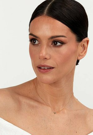 Guldkædetræk med en lille hjertevedhæng, båret på en bar hals. Hudtonen ser strålende ud og har neutral makeup og glat hår.