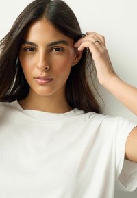 Bílé tričko s volnějším střihem, kulatým výstřihem a ohrnutými rukávy. Modelka má dlouhé vlasy a jemně si jich sahá.