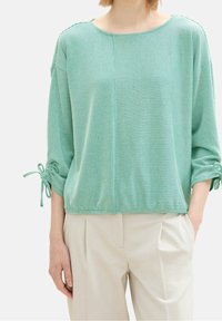 Žena v zeleno-bílém pruhovaném volném topu s 3/4 rukávy svázanými na manžetách, doplněná krémovými kalhotami s vysokým pasem a sklady.