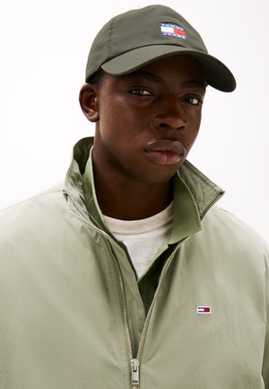 Casquette vert olive avec un écusson logo, associée à une veste zippée vert clair avec un petit logo sur la poitrine et une chemise beige en dessous.