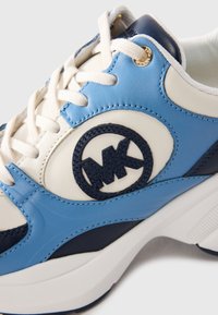 Fehér és kék sneaker krémszínű fűzőkkel, sötétkék kör alakú logóval hímezve az oldalon és aranyszínű fűzőlyukakkal.