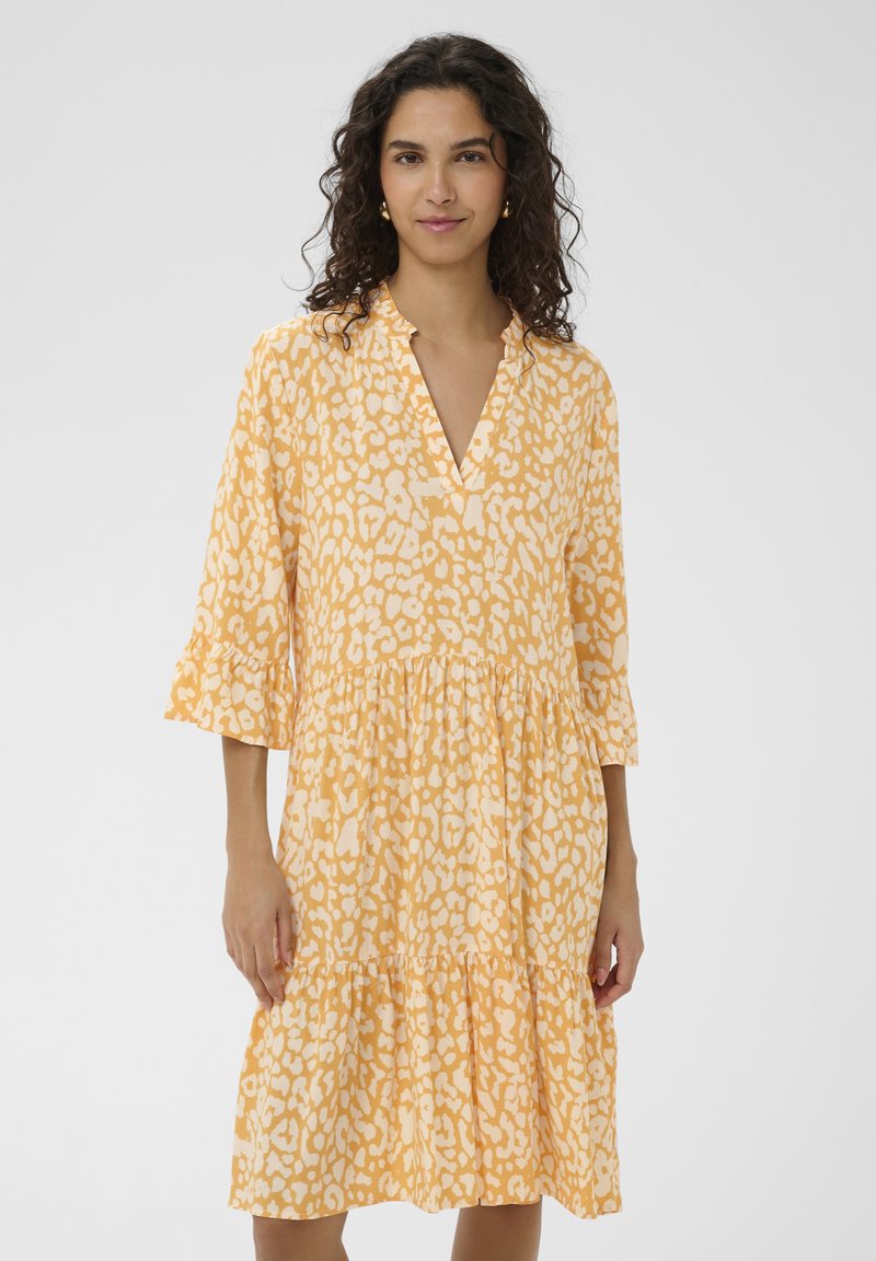Femme aux cheveux bouclés et foncés portant une robe ample jaune et blanche à motifs, manches trois-quarts et col en V, debout devant un fond uni.