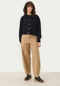 Navy knitted cardigan with a round neckline, button front closure, and two front pockets, paired with tan wide-leg trousers and black shoes.