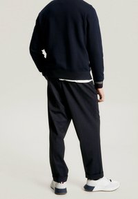 Osoba stojąca tyłem, ubrana w luźne czarne spodnie, granatowy sweter i białe trampki z niebieskimi i czerwonymi detalami na jasnym tle.