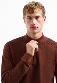 Jeune homme aux cheveux blonds bouclés portant un pull en laine texturé marron, en train de zipper partiellement ou d'ajuster son col, regardant devant lui.