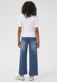 Top blanco de manga corta combinado con pantalones vaqueros de pierna ancha en azul con un lavado desgastado y grandes bolsillos traseros. Zapatillas completan el atuendo.