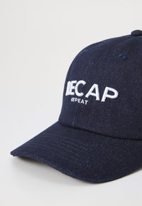 Marineblaue Baseballmütze aus Denim-Material; verfügt über weißen, gestickten Text "RECAP REPEAT" auf der Vorderseite und einem gebogenen Schirm.