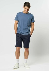 Modrá bavlněná tričko s malým zeleným logem, v kombinaci s tmavě modrými šortkami a bílými sportovními ponožkami. Model má na sobě bílé tenisky a ruce v kapsách.