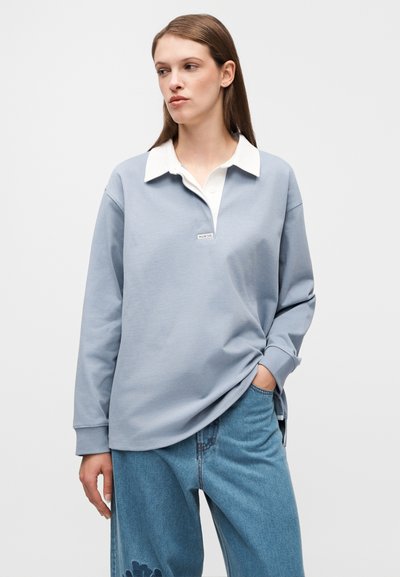 Ανοιχτό μπλε πόλο πουκάμισο με μακριά μανίκια και λευκό γιακά, με ετικέτα στο στήθος και χαλαρή εφαρμογή. Συνδυάζεται με άνετο τζιν παντελόνι.