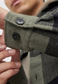 Camisa de cuadros verde con acentos negros, con botones negros en los puños. Primer plano de la textura de la tela y el detalle del botón.
