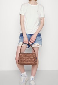 Henkilö, joka on pukeutunut valkoiseen t-paitaan, revittyihin farkkushortseihin ja valkoisiin Adidas-tennareihin, pitää ruskeaa kuvioitua käsilaukkua molemmilla käsillään.