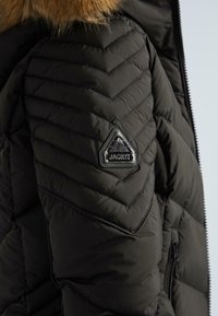 Melna quiltota ziemas jakas piedurkne ar kažoka apmali un trīsstūra metāla logo uzpleca, kurā ir uzraksts "JACK1T".