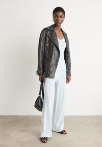 Schwarze Lederbikerjacke, weißes Rippenshirt, hellblaue Schlaghose mit gewelltem Saum, schwarze Handtasche und schwarze Sandalen. Model steht.