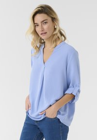 Šviesiai mėlyna V formos blusa su sukryžiuotais rankovėmis ir mygtukų akcentais. Pagaminta iš minkštos medžiagos, turinti laisvą kirpimą ir subtilią tekstūrą.