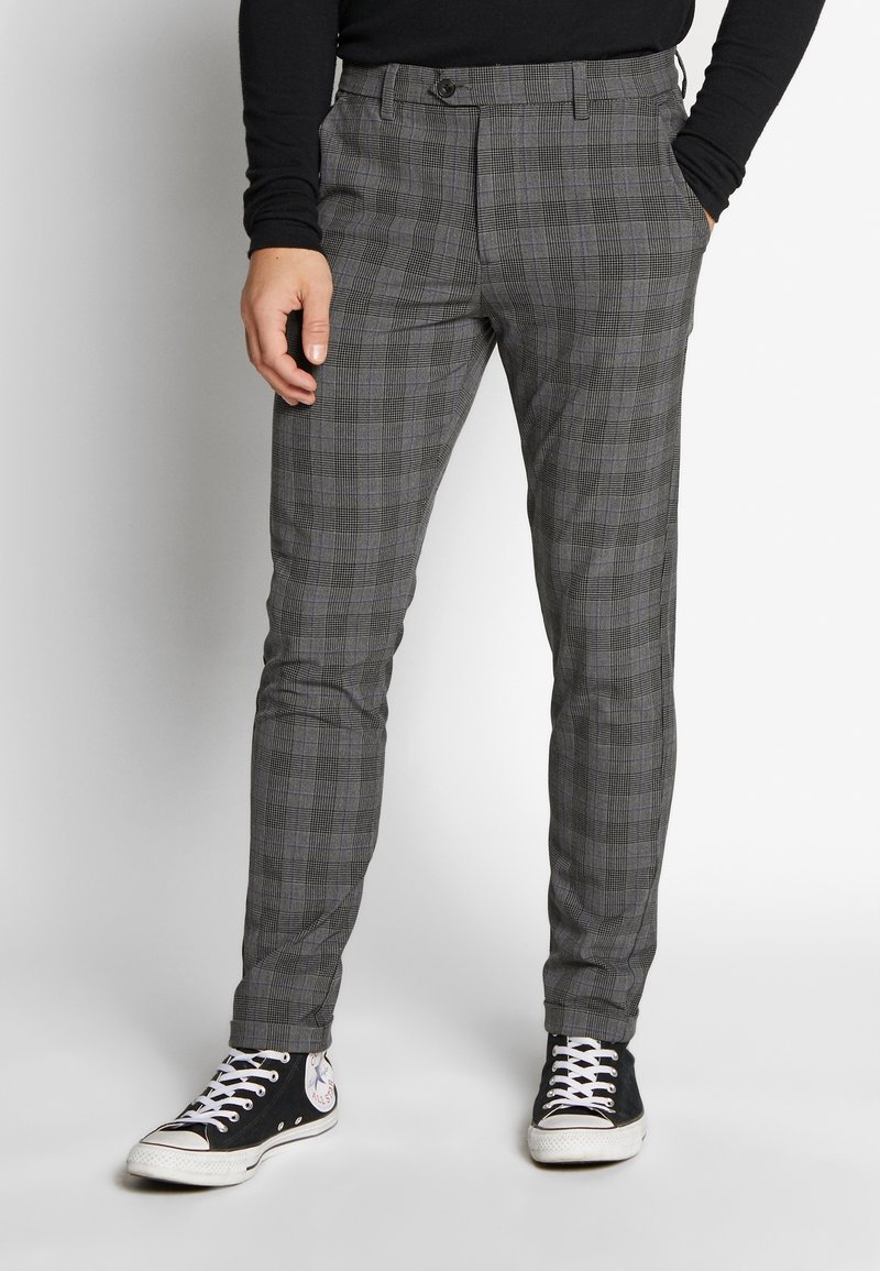 Homme portant un pantalon slim gris à carreaux avec des baskets montantes noires et blanches, debout avec une main dans la poche sur un fond blanc.