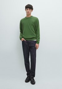 Zielony sweter z okrągłym dekoltem wykonany z dzianiny, zestawiony z ciemnymi jeansami. Model nosi czarne buty i stoi z jedną ręką w kieszeni.