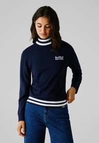 Maglione blu navy con collo alto, rifinito con colletto e orlo a righe bianche, caratterizzato dal logo "Red Bull Racing" in bianco sul petto. Tessuto morbido.