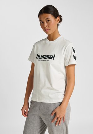 Crème T-shirt met korte mouwen heeft een vet zwart "Hummel" logo en de tekst "sportswear since 1923". Grijze textuurbroek complementeren de outfit.