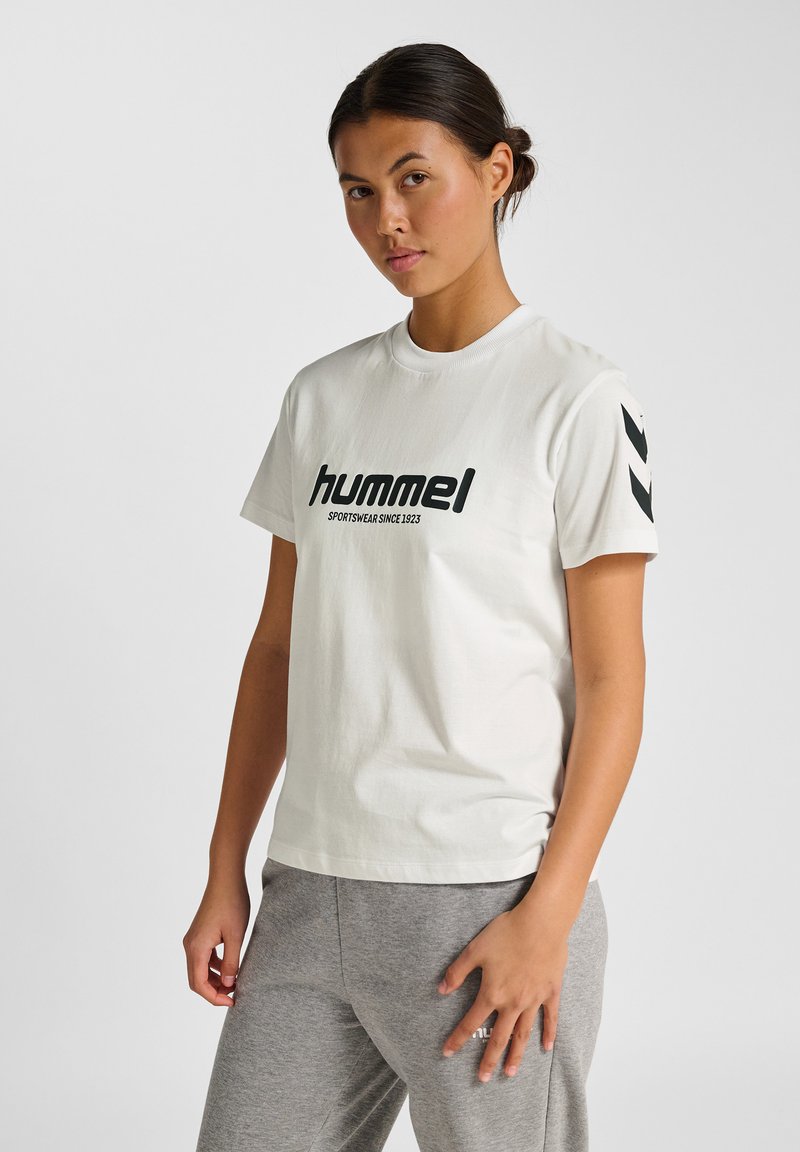 Cremefarvet kortærmet t-shirt har et iøjnefaldende sort "Hummel" logo og teksten "sportswear since 1923". Grå teksturerede bukser supplerer outfittet.
