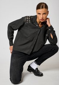 Chemise noire avec des œillets sur les épaules, deux poches avant. Portée avec un jean noir et des mocassins noirs épais. Chaussettes blanches avec des rayures noires.