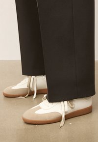 Baltas un beidzs kedas ar gludu ādas augšdaļu, zamša akcentiem un brūnu gumijas zoli, novietotas blakus tumšiem biksēm uz neitrālas grīdas.