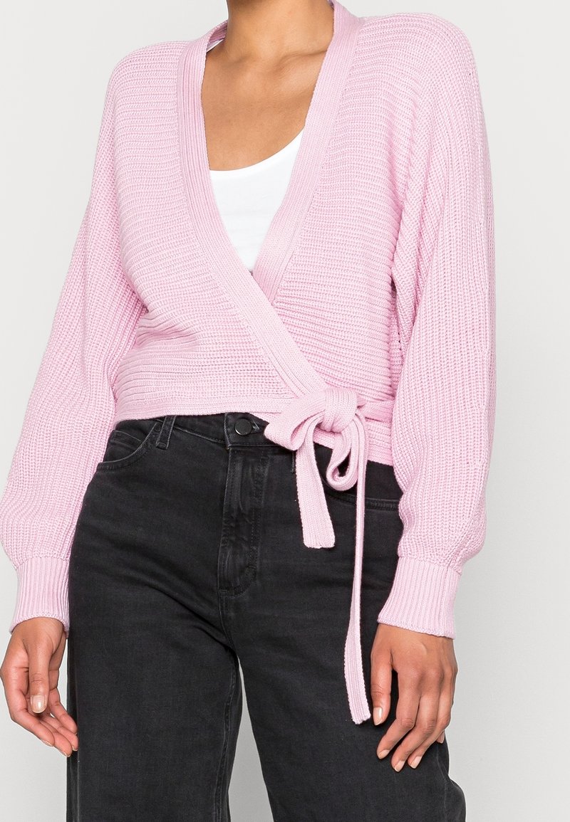 Suéter envolvente de punto rosa con mangas largas, escote en V y cinturón de lazo en la cintura. La textura es acanalada con un acabado suave. Combinado con jeans oscuros.