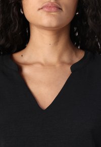 Haut noir à col en V, en tissu léger, avec une texture lisse et une coupe décontractée. Présente un décolleté discret en arrondi et des manches courtes.