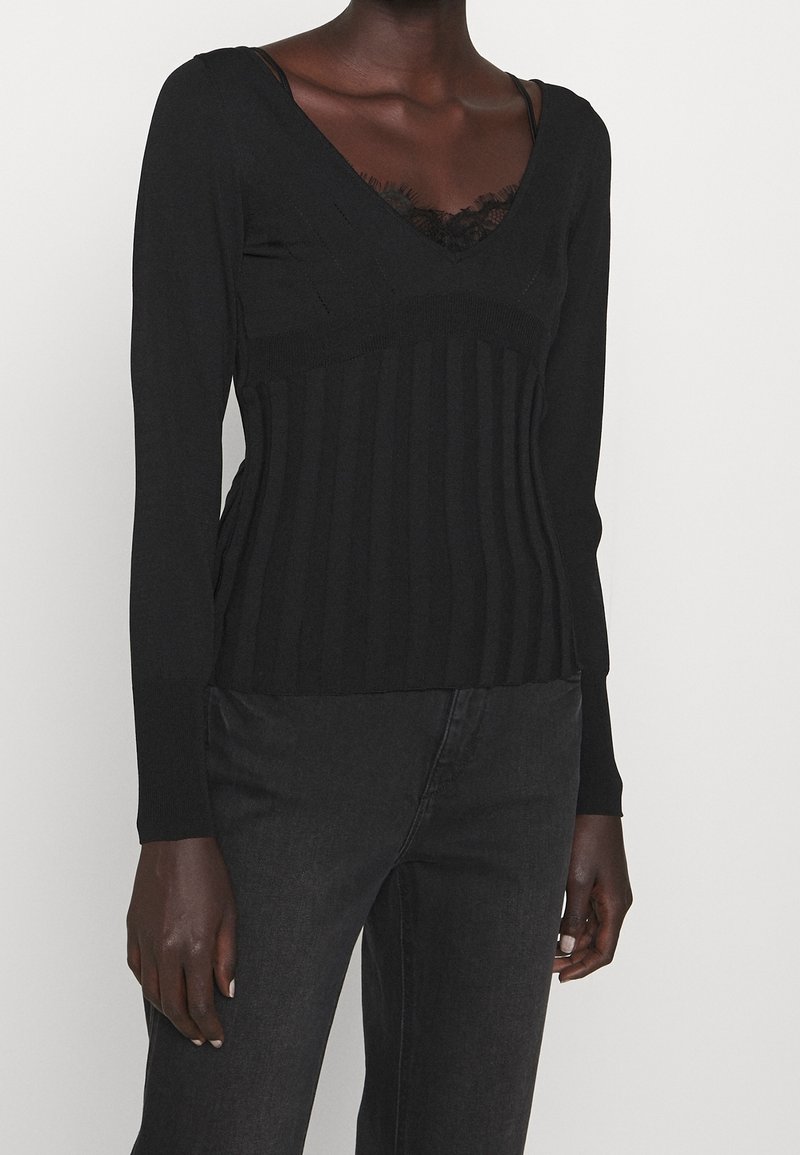 Femme portant un haut noir côtelé à manches longues col en V avec bordure en dentelle, associé à un jean noir, sur un fond uni clair.