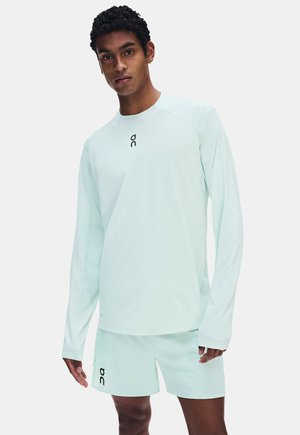 Homme portant une chemise bleu clair à manches longues et un short assorti avec un logo noir, debout devant un fond blanc uni.