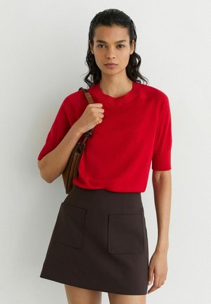 Mujer con un top rojo de manga corta y falda marrón oscuro con bolsillos delanteros, llevando un bolso marrón de hombro sobre un fondo blanco liso.