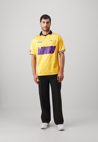 Gelbes Kurzarmtrikot mit schwarzem Kragen, verziert mit lila "LAKERS"-Text; kombiniert mit schwarzen Hosen und weißen Sneakers.