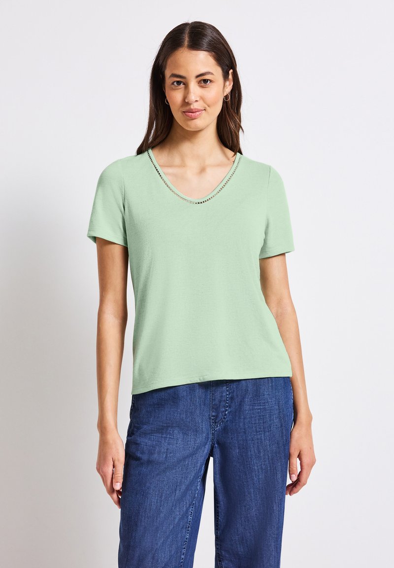 Jasnozielona koszulka z krótkim rękawem i dekoltem w kształcie litery V, ozdobiona dekoracyjną linią małych ćwieków wzdłuż kołnierza, zestawiona z ciemnoniebieskimi dżinsami.