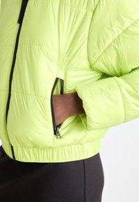 Doudoune verte fluo avec une poche latérale zippée, des poignets et une ceinture élastiques. Texture lisse avec un design matelassé et des accents de fermeture éclair noire.