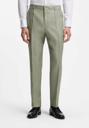 Mand iført lysegrønne skræddersyede bukser, hvid skjorte og sorte formelle sko, stående mod en hvid baggrund.