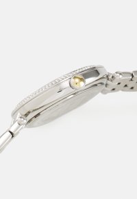 Reloj de metal plateado con una caja redonda, adornado con pequeñas piedras claras a lo largo del bisel y un detalle amarillo en la corona. Pulsera de eslabones de metal.