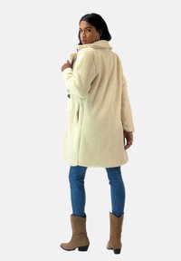 Abrigo de piel sintética de color crema con cuello, dos botones, bolsillos laterales y una superficie texturizada, combinado con jeans azules y botas marrones.